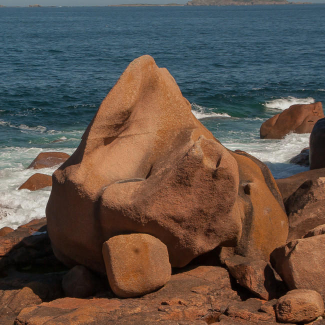 Ein großer, rötlicher Granitblock steht an einer Felsenküste in der Brandung.